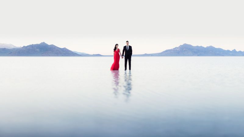 two people stand, center frame, surrounded by water and mountains in a stitched panorama